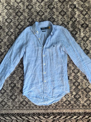 Blå linne skjorta Ralph Lauren  - Denna skjorta från Ralph Lauren är i tyget linne som är perfekt till sommaren. Den är inte använd speciellt mycket och är i fint skick. Modell och storlek ör custom fit i storlek xs den sitter som en slim fit. Jag kan ungefär ha den och är 182. Ny pris 1500 mitt pris 750