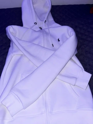 Vit hoodie från Polo Ralph Lauren - Säljer en clean vit hoodie från Polo Ralph Lauren med dragkedja framtill och klassisk huva med snören. Ikoniska lilla loggan broderad på bröstet. Hoodien har kängurufickor och ribbade muddar. Perfekt för en chill och stilren look.