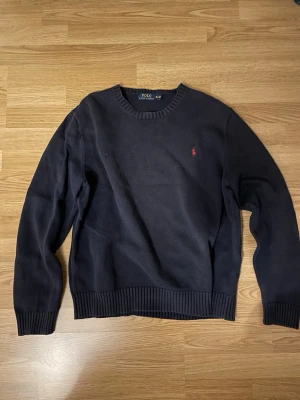 Ralph Lauren tröja - Klassisk mörkblå stickad tröja från Polo Ralph Lauren med rund halsringning och röd broderad logga på bröstet. Tröjan har ribbstickade muddar vid ärmslut och nederkant. Perfekt till lager på lager och enkel att matcha. Storlek XL men passar M/L