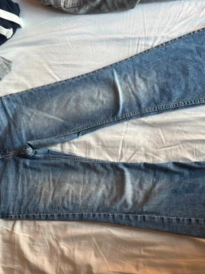 Blå jeans i rak modell - Säljer ett par klassiska blå jeans i rak modell med snygga slitningar och ljusare partier på låren. Jeansen har normal midja och är tillverkade i denim. Perfekta för en avslappnad och trendig look.