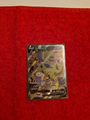 Tyranitar V Pokémonkort - Säljer ett Tyranitar V Pokémonkort med färgstark illustration och holografisk yta. Kortet har motiv av Tyranitar i en skogsmiljö och är från Single Strike-serien. Perfekt för samlare eller dig som vill uppgradera din kortlek.
