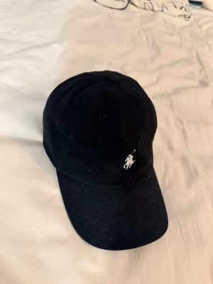 Svart keps från Polo Ralph Lauren - Säljer en svart keps från Polo Ralph Lauren med klassisk böjd skärm och broderad vit logga framtill. Justerbart spänne bak och 'POLO' broderat i vitt. I bomull