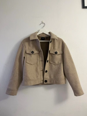 Beige overshirt från Vero Moda - Stilren beige overshirt från Vero Moda i ullmix med klassisk krage, stora bröstfickor med knappar och svarta knappar framtill. Rak passform och långa ärmar. Perfekt lager på lager-plagg för en trendig look.