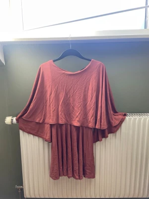 Röd/brun omlottklänning med volang - Säljer en flowy brun/röd klänning med omlottkänsla och volangdetaljer. Klänningen har vid passform, rund halsringning och ärmlös design med cape-liknande överdel. Tillverkad i mjukt, tunt tyg som faller snyggt.