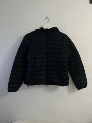 Svart pufferjacka med huva - Säljer en svart pufferjacka med huva och dragkedja framtill. Jackan har två sidofickor med dragkedja och quiltad design. Perfekt för kalla dagar och riktigt skön att bära. Passar dig som gillar en enkel och stilren look.