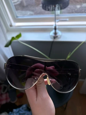 Vinted Gucci sheild solglasögon  - Snygga solglasögon med stora, svarta pilotformade linser och guldfärgade metallbågar. Glasögonen har en modern och trendig look med dubbla bryggor och justerbara näskuddar för extra komfort.