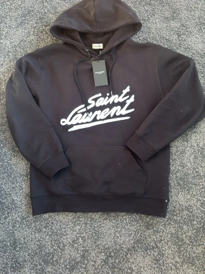 Saint Laurent hoodie - Svart hoodie från Saint Laurent med stor vit logga i retrostil på bröstet. Klassisk huva med dragsko och känguruficka framtill. Tillverkad i mjukt material som känns skönt mot huden. Perfekt för dig som vill ha en stilren och trendig look.