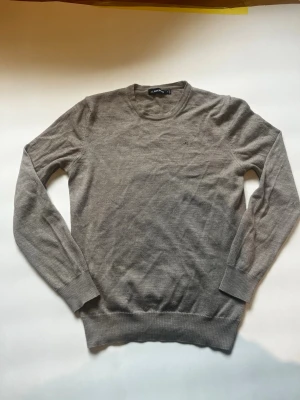 Grå stickad tröja från J.Lindeberg - Klassisk grå stickad tröja från J.Lindeberg med rund halsringning och ribbade muddar vid ärmslut och nederkant. Tröjan har en diskret logga på bröstet och är tillverkad i ett tunt, mjukt material som passar perfekt till lager-på-lager. Något litet hål finns i ärmarna annars är den fin, storleken är M men passar den som bär S