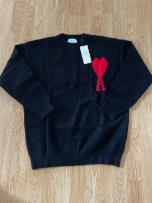 Ami Paris tröja - Snygg svart stickad tröja från Ami Paris med röd hjärtlogga och bokstaven A på bröstet. Tröjan har rund halsringning och ribbade muddar vid ärmslut och nederkant. Perfekt för dig som gillar stilrena plagg med en twist.