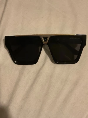 Svarta Louis Vuitton solglasögon - Snygga svarta solglasögon från Louis Vuitton med rektangulär form och gulddetaljer. Glasögonen har en bred båge och tydlig LV-logga på sidan. Perfekt accessoar för dig som vill sticka ut med en exklusiv look. LV glasögon använt ett fåtal gånger.