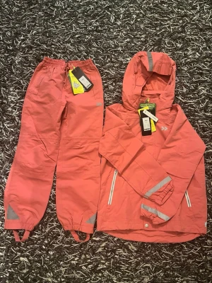 Rosa vindjacka med huva från Didriksons - Snygg rosa vindjacka från Didriksons med huva och reflexdetaljer på ärmar och rygg. Jackan har dragkedja framtill, två fickor med vita dragkedjor och justerbara muddar. Materialet är slitstarkt och vattenavvisande, perfekt för blåsiga dagar.