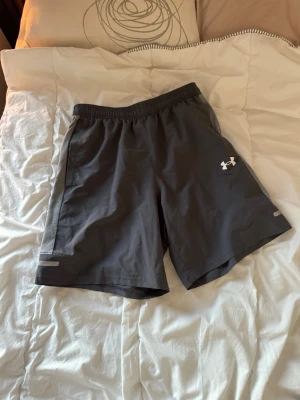 Grå träningsshorts Under Armour - Säljer ett par mörkgrå träningsshorts från Under Armour med elastisk midja och reflekterande detaljer. Shortsen har en sportig look och är tillverkade i lätt syntetmaterial som andas, perfekta för aktiva dagar. Storlek S