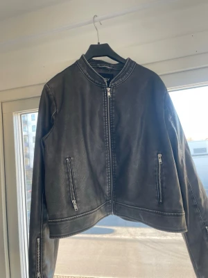 Grå skinnjacka från ONLY - Snygg svart skinnjacka från ONLY med raka linjer och silverfärgade dragkedjor på fickorna och framtill. Jackan har en minimalistisk design utan krage och är helfodrad på insidan. Perfekt för dig som gillar en stilren look.