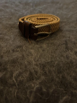 La Boucle flätat bälte - Snyggt beige flätat bälte med bruna skinn-detaljer och silverfärgat spänne. Bältet har en klassisk look och passar perfekt till jeans eller chinos. Flätad design gör det enkelt att justera längden. Liten röd detalj vid spetsen för extra stil.