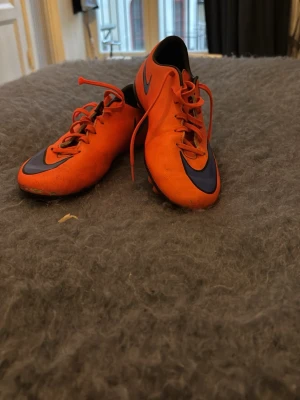 Nike orange fotbollsskor med dobbar - Säljer ett par grymma orangea fotbollsskor från Nike med klassisk svart swoosh på sidan. Skorna har snörning och fasta dobbar för bra grepp på planen. Tillverkade i syntetmaterial som ger lätt känsla och bra bollkontroll. Perfekta för dig som vill sticka ut på matchen.