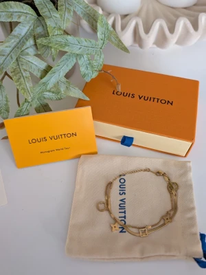 Iouis vuitton armband  - Elegant dubbelradigt armband i guldfärgad metall med tillhörande originalförpackning. Design: Armbandet består av två olika kedjor – en klassisk nätt länk och en plattare "snake"-kedja. Detaljer: Dekorerat med flera hängande berlocker i form av stiliserade fyrbladiga blommor och geometriska figurer. Tillbehör: Säljes komplett med en orange signaturask, en ljus dammpåse i tyg samt ett tillhörande infokort ("Monogram World Tour"). Stängning: Justerbar längd med klolås för optimal passform.