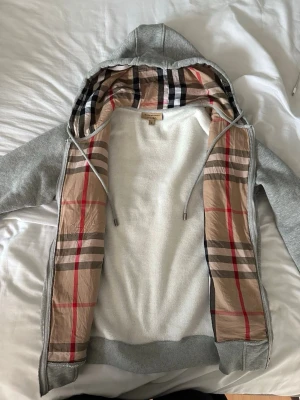 Grå hoodie från Burberry med mönstrad kant - Säljer en grå hoodie från Burberry med dragkedja och den klassiska beigea Burberry-rutiga kanten på insidan samt i huvan. Hoodien har dragsko i huvan och ribbade muddar. Materialet är mjukt och bekvämt, perfekt för en avslappnad men ändå stilren look.