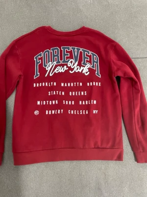 Röd sweatshirt Forever New York - Snygg röd sweatshirt med tryck på ryggen där det står 'Forever New York' och namn på olika stadsdelar i New York. Tröjan har rund halsringning, långa ärmar och ribbade muddar. Perfekt för dig som gillar streetwear och vill sticka ut med ett coolt tryck.