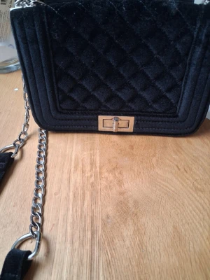 Svart quiltad axelremsväska från Ur&Penn - Säljer en svart handväska med quiltat mönster och kedjerem. Väskan har en rektangulär form, guldfärgat vridlås och är gjord i ett mjukt sammetstyg. Perfekt för dig som gillar en stilren och trendig look.
