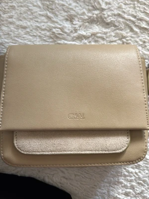 Beige axelväska från C.W. - Snygg beige axelväska från C.W. i stilren, fyrkantig modell. Väskan har justerbar axelrem och en unik detalj med en ficka i beige tyg på framsidan. Tillverkad i slätt konstläder med silverfärgade metalldetaljer. Perfekt för dig som gillar minimalistisk design.