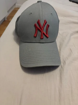 Grå New Era NY keps - Snygg grå keps från New Era med röd broderad NY-logga framtill. Modellen är 39THIRTY och har böjd skärm samt stretchig passform. Tillverkad i bomullsmix och officiell MLB-merchandise. Perfekt för dig som gillar streetwear och sportig stil.