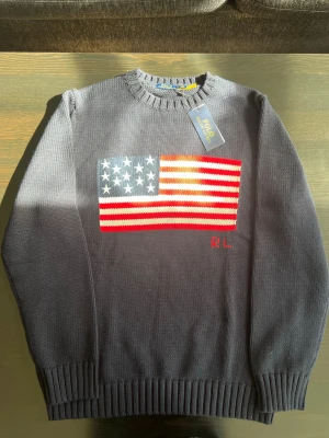 Mörkblå stickad tröja Polo Ralph Lauren - Mörkblå stickad tröja från Polo Ralph Lauren med amerikansk flagga broderad på bröstet och RL-broderi i rött. Tröjan har ribbstickade muddar vid ärmslut och nederkant samt rund halsringning. Perfekt för dig som gillar klassisk preppy stil.