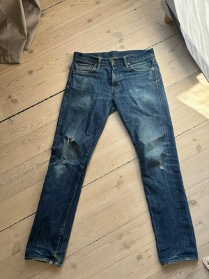 Selvedge levi jeans 511 - Säljer ett par blå selvedge levi jeans med raka ben och slitningar på knäna. Jeansen har klassisk femficksdesign, knappgylf och kontrastsömmar. Perfekta för en avslappnad och trendig look.