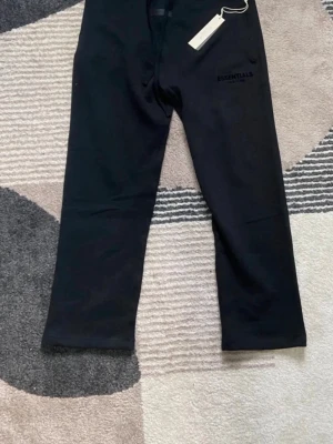 Svarta Essentials mjukisbyxor - Snygga svarta mjukisbyxor från Essentials med diskret logga på benet. Byxorna har resår i midjan och raka ben. Tillverkade i mjukt bomullsmaterial som känns skönt mot huden. Perfekta för en avslappnad och trendig look.