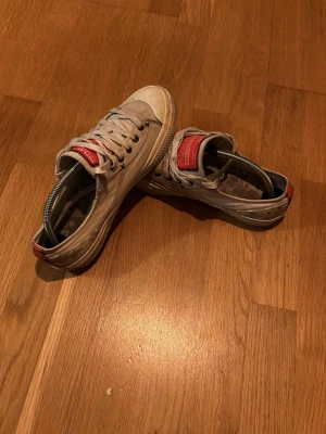 Vita Replay sneakers med röd detalj - Säljer ett par vita låga sneakers från Replay med röda detaljer på plösen och hälen. Skorna har vita snören och en sula i gummi. Insidan har ett grått camomönster. Perfekta för dig som gillar en clean och stilren look. Skorna är ganska välanvända och kan behöva lite kärlek men extremt unika och snygga.