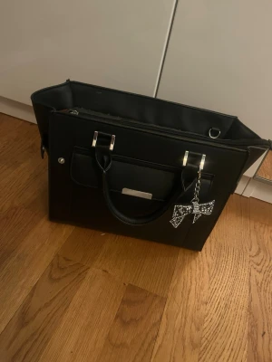 Svart väska - Snygg svart med både handtag och justerbar axelrem. Väskan har silverfärgade detaljer, en ficka framtill och en dekorativ nyckelring med strass. Insidan är fodrad i brunt tyg och har flera fack för organisering. Köpte för 700kr