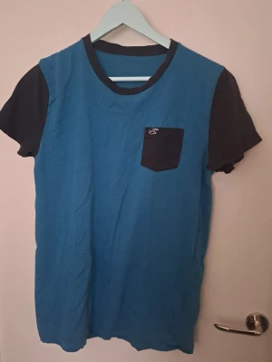 Blå t-shirt med svart ficka Hollister - Snygg blå t-shirt från Hollister med svarta ärmar, svart bröstficka och svart krage. Fickan har en liten broderad logga. Perfekt till jeans eller shorts för en avslappnad look. Tror det är en L i Storlek. 