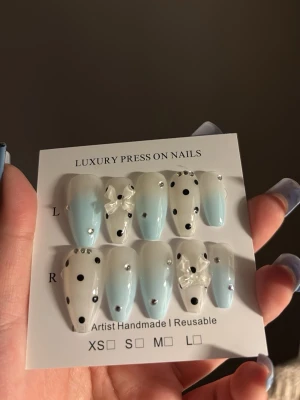 Blå press-on naglar med prickar & rosetter - Snygga press-on naglar i ljusblått och vitt med svarta prickar, små silverfärgade stenar och söta 3D-rosetter. Naglarna har en elegant almondform och är handgjorda. Perfekt för dig som vill ha en unik och trendig look.