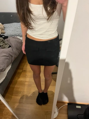 Pull&bear svart kortkjol med fickdetaljer - Säljer en svart kortkjol med två dekorativa fickor framtill, med dragkedja på sidan. Kjolen har två målarfärg fläckar på sidan, se bild 3