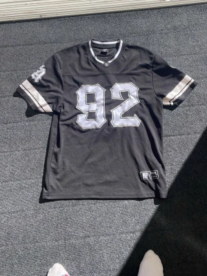 Svart LA t-shirt med nummer 92 - Säljer en svart t-shirt i sportig stil med stora vita siffror 92 på både fram- och baksidan samt texten 'LOS ANGELES' på ryggen. T-shirten har vita och grå ränder på ärmarna och en rund halsringning. Perfekt för dig som gillar streetwear och amerikansk sportstil.