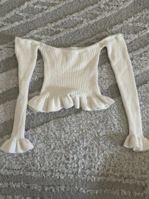 Vit ribbad topp med volang - Säljer en vit ribbad topp med långa ärmar och volangdetaljer ärmslut och nederkant. Toppen har en figurnära passform och offshoulder-design. 