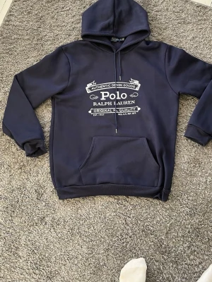 Marinblå hoodie Polo Ralph Lauren - Snygg marinblå hoodie från Polo Ralph Lauren med vit tryckt logga och text på bröstet. Klassisk känguruficka framtill, justerbar huva med dragsko och ribbade muddar. Perfekt för en avslappnad och stilren look.