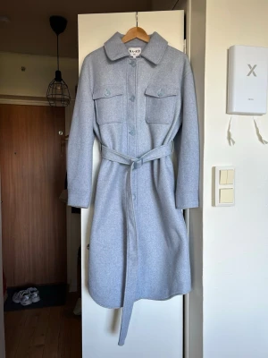 Ljusblå kappa från NA-KD - Snygg ljusblå kappa från NA-KD med knappar framtill, två stora bröstfickor och ett matchande knytskärp i midjan. Klassisk krage och rak passform som ger en stilren look. Perfekt för kyliga dagar och enkel att styla till olika outfits.