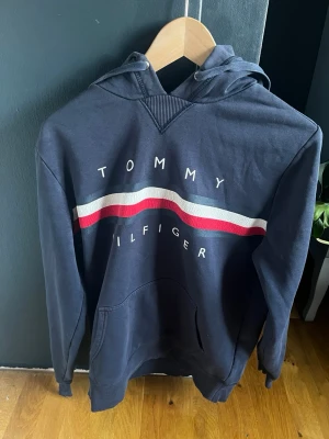 Marinblå hoodie Tommy Hilfiger - Snygg marinblå hoodie från Tommy Hilfiger med klassisk röd och vit rand över bröstet (lite sletet) och stort tryck med loggan. Hoodien har huva och känguruficka framtill. Tillverkad i mjukt bomullsmaterial för skön känsla och avslappnad stil.