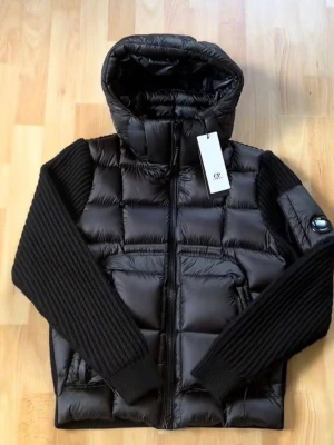 Svart pufferjacka från C.P. Company - Snygg svart pufferjacka från C.P. Company med glansig finish, stickade ärmar och huva. Jackan har två framfickor med dragkedja och en ikonisk linsdetalj på ena ärmen. Perfekt för kalla dagar och riktigt trendig look.