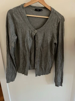 Grå kofta  - En enkel och stilren grå kofta från Fransa med v-ringning och knäppning framtill. Koftan har långa ärmar och ribbade muddar vid ärmslut och nederkant. 