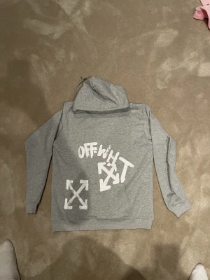 Off White hoodie  - Snygg grå hoodie från Off-White med vita tryck både fram och bak. Klassisk huva med dragsko och ribbade muddar. Stora grafiska detaljer och logga ger en streetwear vibe. Perfekt för dig som gillar trendiga och bekväma plagg. Stl L passar M
