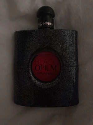 Yves Saint Laurent Black Opium parfym - Black Opium från Yves Saint Laurent är en ikonisk parfym i en svart, glittrig flaska med rund röd detalj i mitten. Flaskan har en elegant och modern design med tydlig logga på korken. Förpackningen är svart med silverdetaljer och YSL-logga.