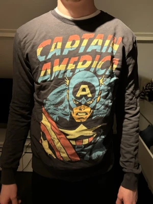 Captain America sweatshirt - Cool grå sweatshirt med stort Captain America-tryck i färgerna blå, gul, röd och vit på framsidan. Tröjan har rund halsringning, långa ärmar och ribbade muddar vid ärmslut och nederkant. Perfekt för Marvel-fans som vill sticka ut.