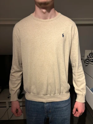 Beige tröja från Ralph Lauren - Klassisk beige tröja från Ralph Lauren med rund halsringning och diskret blå logga broderad på bröstet. Tröjan har långa ärmar och ribbade muddar vid ärmslut och nederkant. Perfekt till jeans för en clean och stilren look. Slimfit 