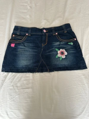 Mörk jeanskjol med broderier - Säljer en mörkblå jeanskjol med broderade blommor, regnbåge och fjärilar. Kjolen har fem fickor, rosa detaljer och rå kant nedtill för en avslappnad look. Perfekt för dig som vill ha en unik och färgglad denimkjol.