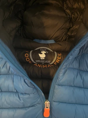 Blå dunjacka från Save The Duck - Snygg blå dunjacka från Save The Duck i ultralätt material. Jackan är 100% animal free och har en vadderad design med dragkedja framtill samt en orange logga på ärmen. Perfekt för kalla vinterdagar och riktigt skön att bära.