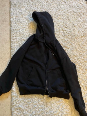 Svart zip hoodie med fickor - En klassisk svart hoodie med dragkedja framtill och två stora magfickor. Hoodien har justerbar huva med snören och ribbade muddar vid ärmslut och nederkant. Perfekt för en avslappnad och stilren look. Materialet är mjukt och bekvämt.