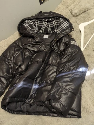 Svart dunjacka från Burberry - Säljer en svart dunjacka från Burberry med klassiskt rutigt foder i huvan och tryck på ärmen. Jackan har dragkedja, knappar och en stor huva. Perfekt för kalla vinterdagar och riktigt snygg streetstil.