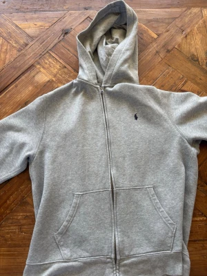 Grå zip hoodie från Polo Ralph Lauren - Snygg grå hoodie med dragkedja från Polo Ralph Lauren. Klassisk design med huva, två stora fickor fram och den ikoniska lilla loggan broderad på bröstet. Tillverkad i mjuk bomullsblandning som är perfekt för chill dagar eller när du vill ha en clean look. Storlek 170cm