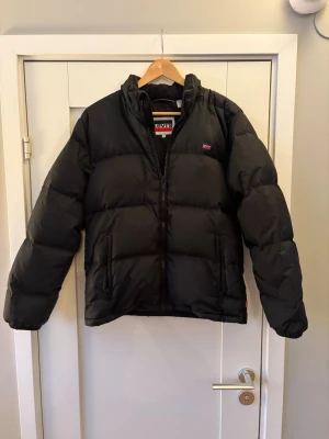 Svart dunjacka från Levis - Snygg och varm svart dunjacka från levis, Tilverkad i 100% dun. Nypris 2799kr säljer den nu för endast 349kr. Finns en liten skråma på ryggen annars som ny i skick.